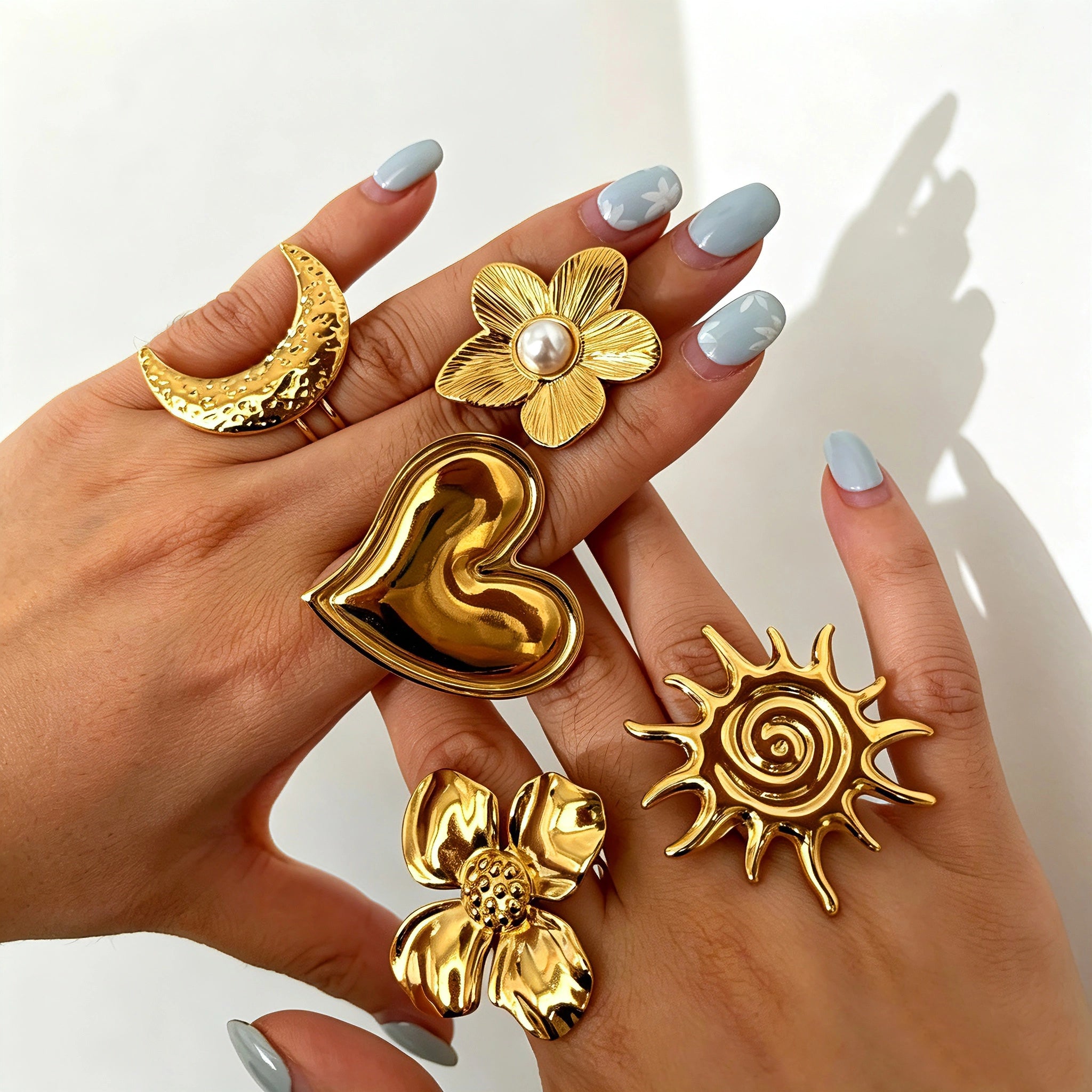 This 18K gold‑plated stainless steel ring set features bold sculpted designs of flowers, a puffy heart, swirling sun, and hammered crescent moon. Some flower rings are accented with a lustrous faux pearl for extra vintage charm. These open adjustable statement rings deliver a chunky retro aesthetic for everyday and special‑occasion styling.