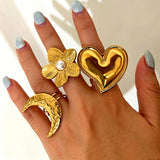 This 18K gold‑plated stainless steel ring set features bold sculpted designs of flowers, a puffy heart, swirling sun, and hammered crescent moon. Some flower rings are accented with a lustrous faux pearl for extra vintage charm. These open adjustable statement rings deliver a chunky retro aesthetic for everyday and special‑occasion styling.