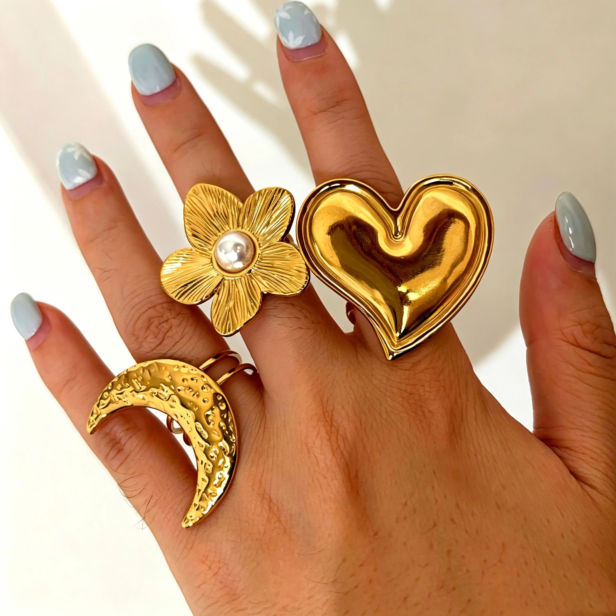 This 18K gold‑plated stainless steel ring set features bold sculpted designs of flowers, a puffy heart, swirling sun, and hammered crescent moon. Some flower rings are accented with a lustrous faux pearl for extra vintage charm. These open adjustable statement rings deliver a chunky retro aesthetic for everyday and special‑occasion styling.