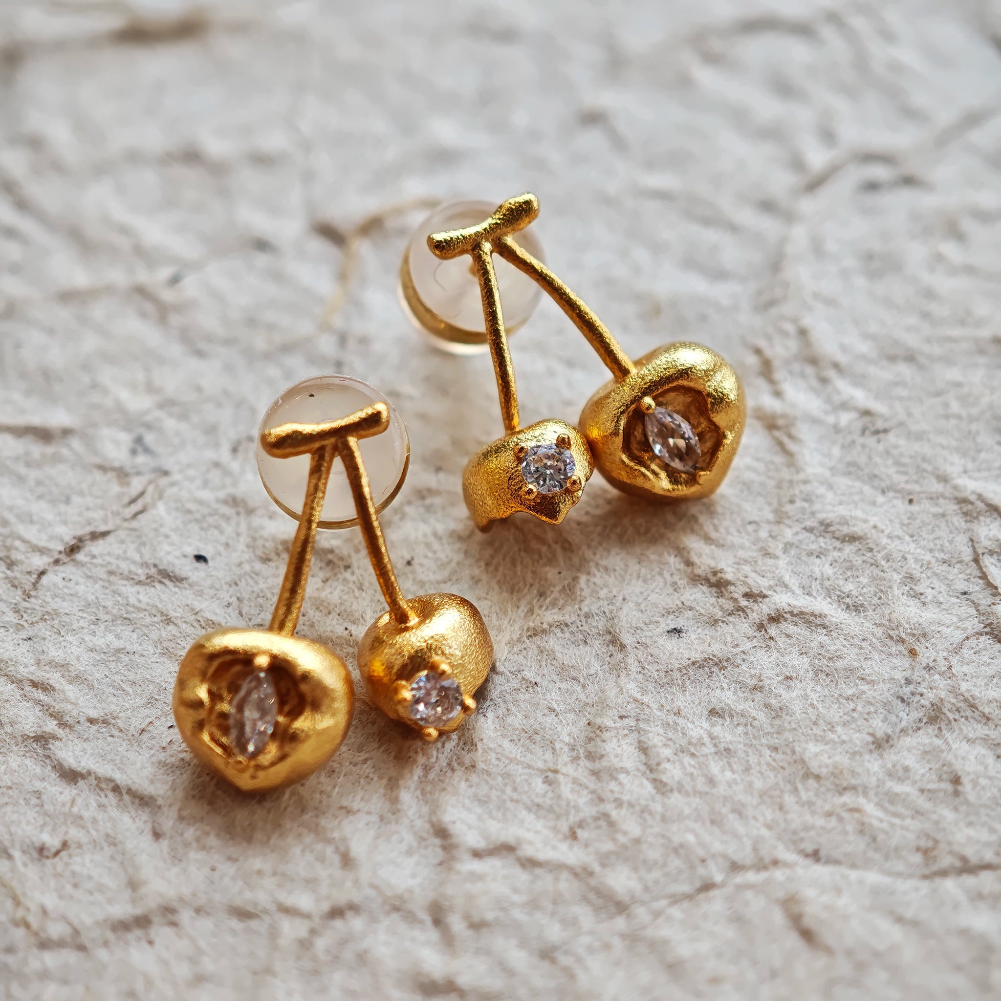 These cherry-shaped stud earrings feature a luxurious matte sandblasted finish, crafted from thick-plated real gold over 925 sterling silver posts. Each earring is designed as a pair of cherries, adorned with a marquise-cut and a round-cut sparkling clear stone, adding a delicate, eye-catching sparkle. The playful, organic cherry silhouette and premium gold plating make them a chic, elegant accessory for everyday wear.