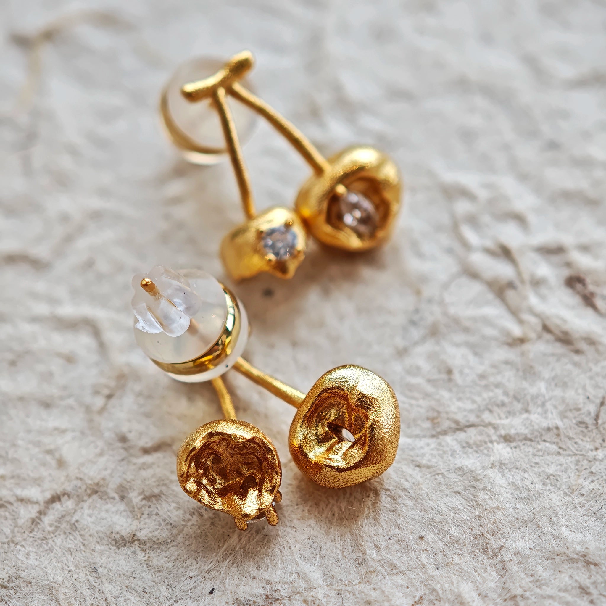 These cherry-shaped stud earrings feature a luxurious matte sandblasted finish, crafted from thick-plated real gold over 925 sterling silver posts. Each earring is designed as a pair of cherries, adorned with a marquise-cut and a round-cut sparkling clear stone, adding a delicate, eye-catching sparkle. The playful, organic cherry silhouette and premium gold plating make them a chic, elegant accessory for everyday wear.