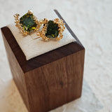 These stud earrings feature cushion-cut olive-green cubic zirconia stones, each encircled by delicate, nature-inspired gold floral and vine detailing. The thick 18K gold plating lends a rich, warm luster, contrasting beautifully with the deep green cubic zirconia to create an elegant, botanical aesthetic.
