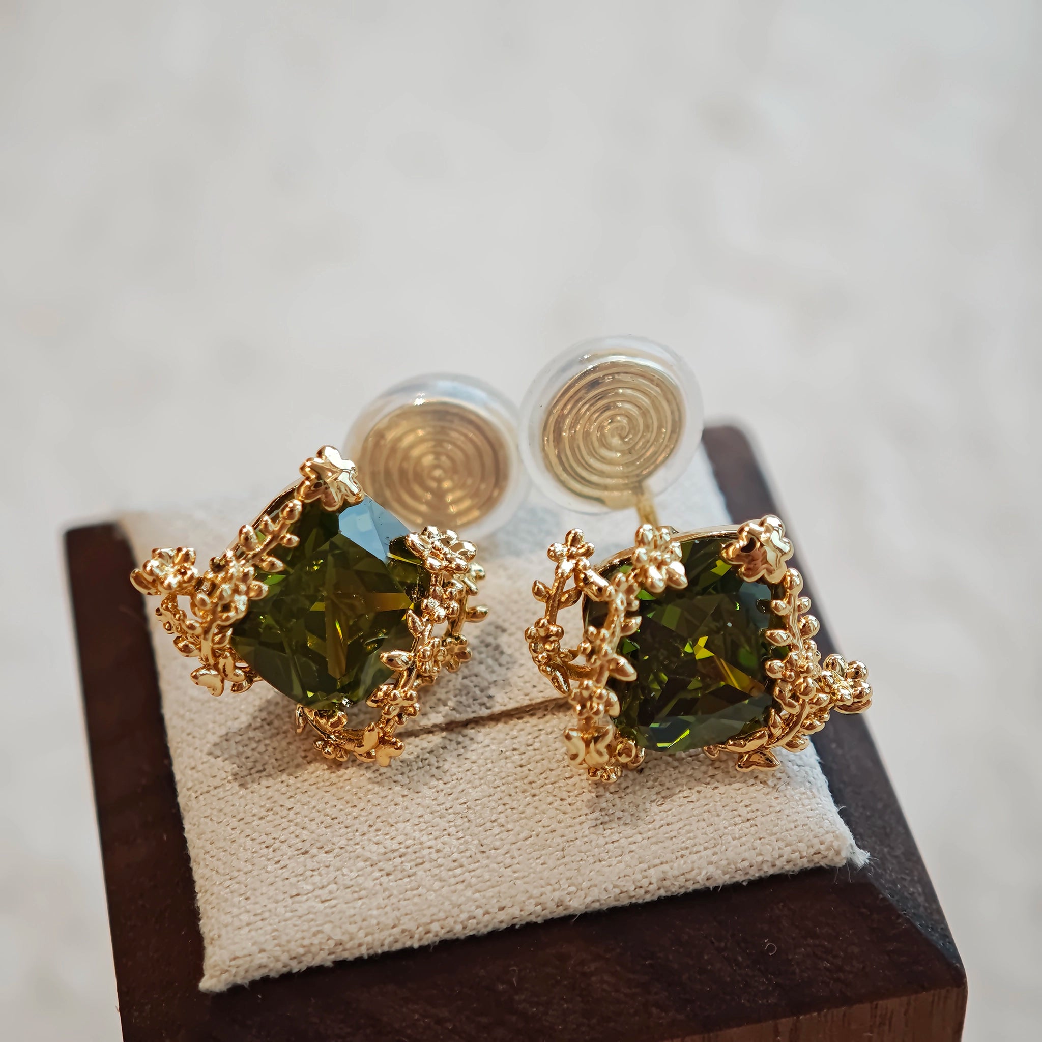 These stud earrings feature cushion-cut olive-green cubic zirconia stones, each encircled by delicate, nature-inspired gold floral and vine detailing. The thick 18K gold plating lends a rich, warm luster, contrasting beautifully with the deep green cubic zirconia to create an elegant, botanical aesthetic.