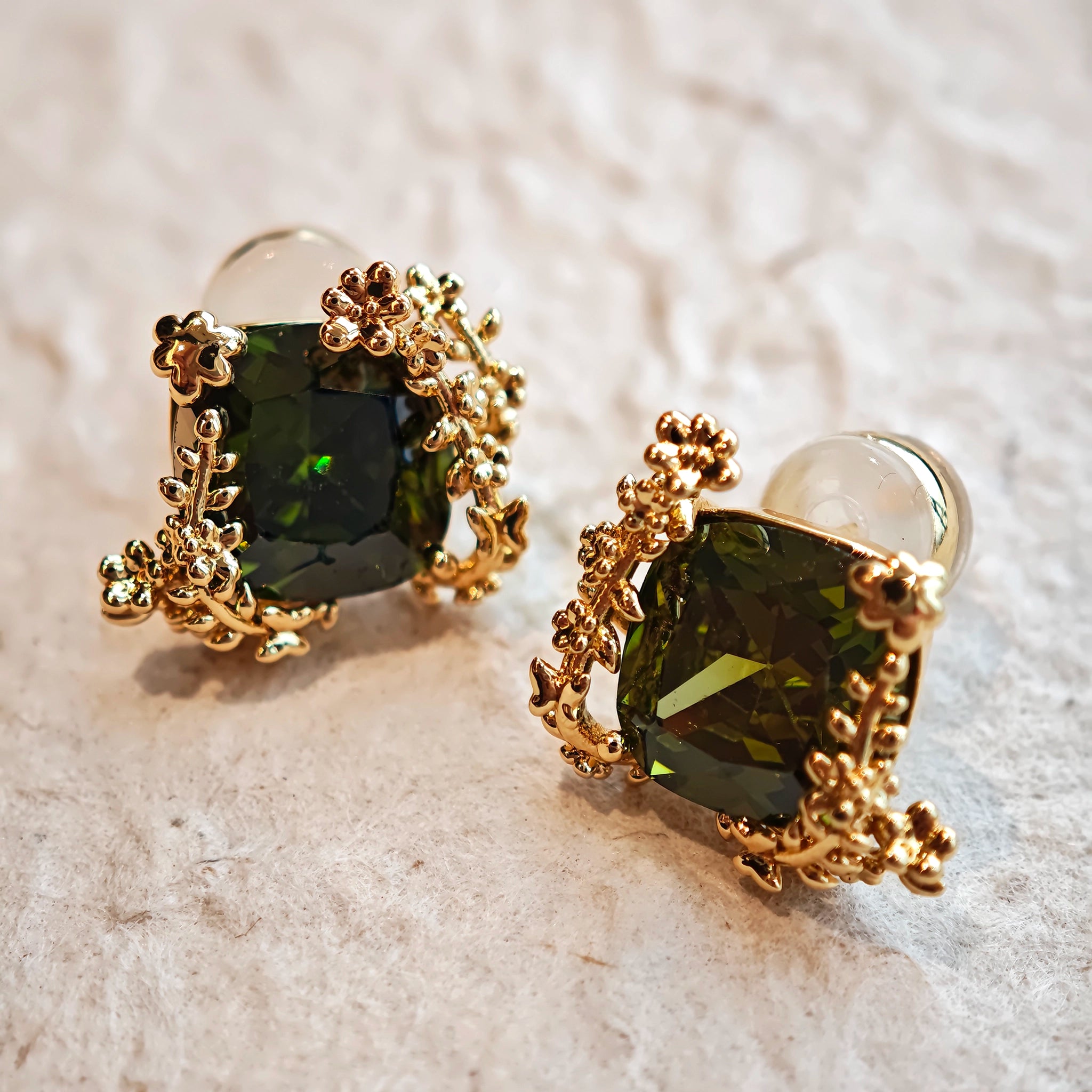 These stud earrings feature cushion-cut olive-green cubic zirconia stones, each encircled by delicate, nature-inspired gold floral and vine detailing. The thick 18K gold plating lends a rich, warm luster, contrasting beautifully with the deep green cubic zirconia to create an elegant, botanical aesthetic.