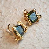 These stud earrings feature cushion-cut olive-green cubic zirconia stones, each encircled by delicate, nature-inspired gold floral and vine detailing. The thick 18K gold plating lends a rich, warm luster, contrasting beautifully with the deep green cubic zirconia to create an elegant, botanical aesthetic.