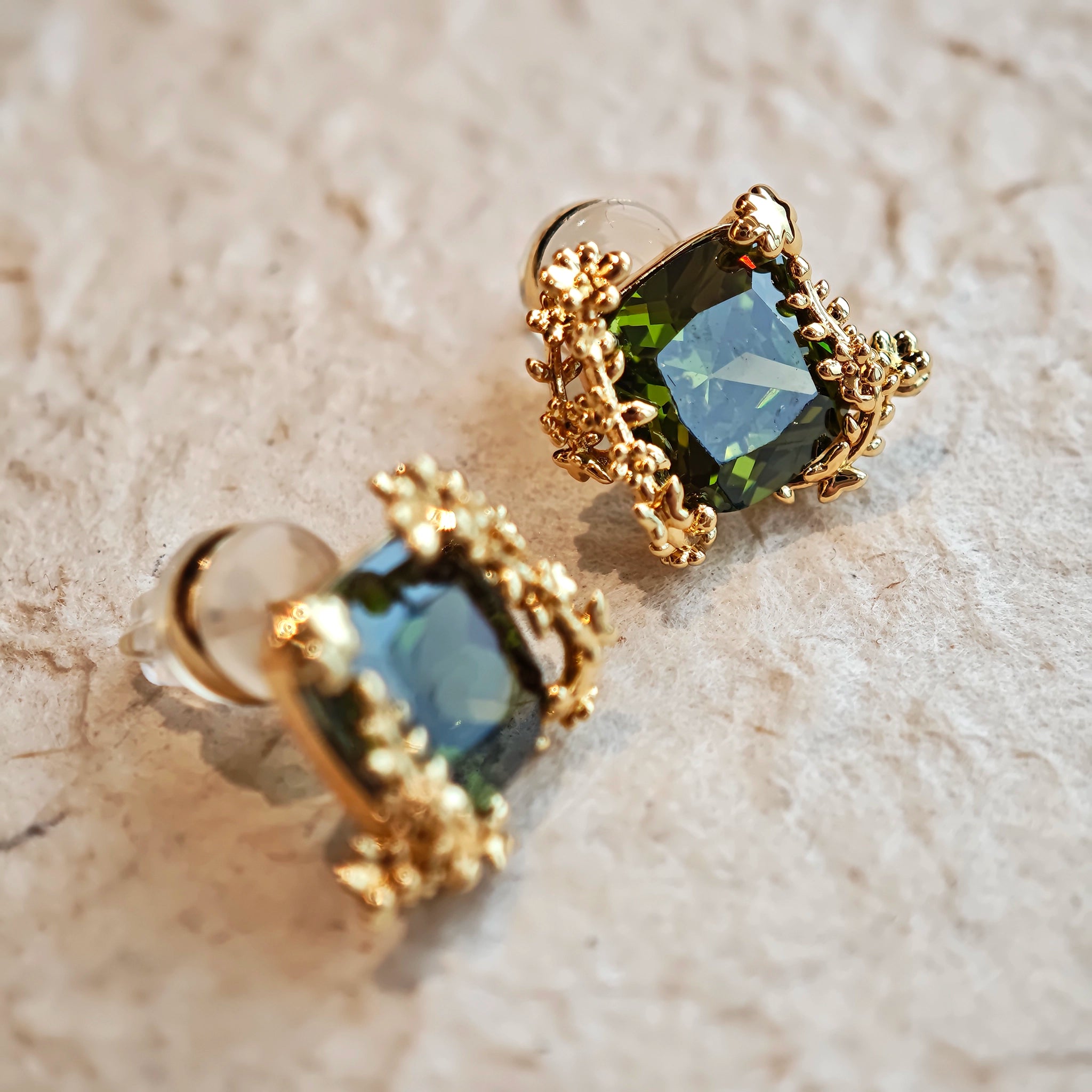 These stud earrings feature cushion-cut olive-green cubic zirconia stones, each encircled by delicate, nature-inspired gold floral and vine detailing. The thick 18K gold plating lends a rich, warm luster, contrasting beautifully with the deep green cubic zirconia to create an elegant, botanical aesthetic.