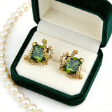 These stud earrings feature cushion-cut olive-green cubic zirconia stones, each encircled by delicate, nature-inspired gold floral and vine detailing. The thick 18K gold plating lends a rich, warm luster, contrasting beautifully with the deep green cubic zirconia to create an elegant, botanical aesthetic.