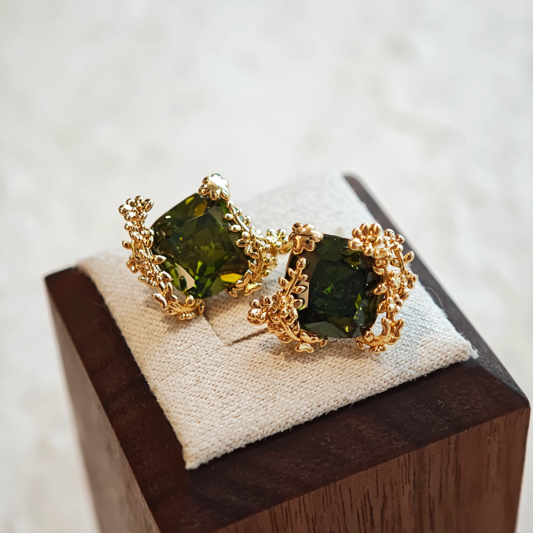 These stud earrings feature cushion-cut olive-green cubic zirconia stones, each encircled by delicate, nature-inspired gold floral and vine detailing. The thick 18K gold plating lends a rich, warm luster, contrasting beautifully with the deep green cubic zirconia to create an elegant, botanical aesthetic.
