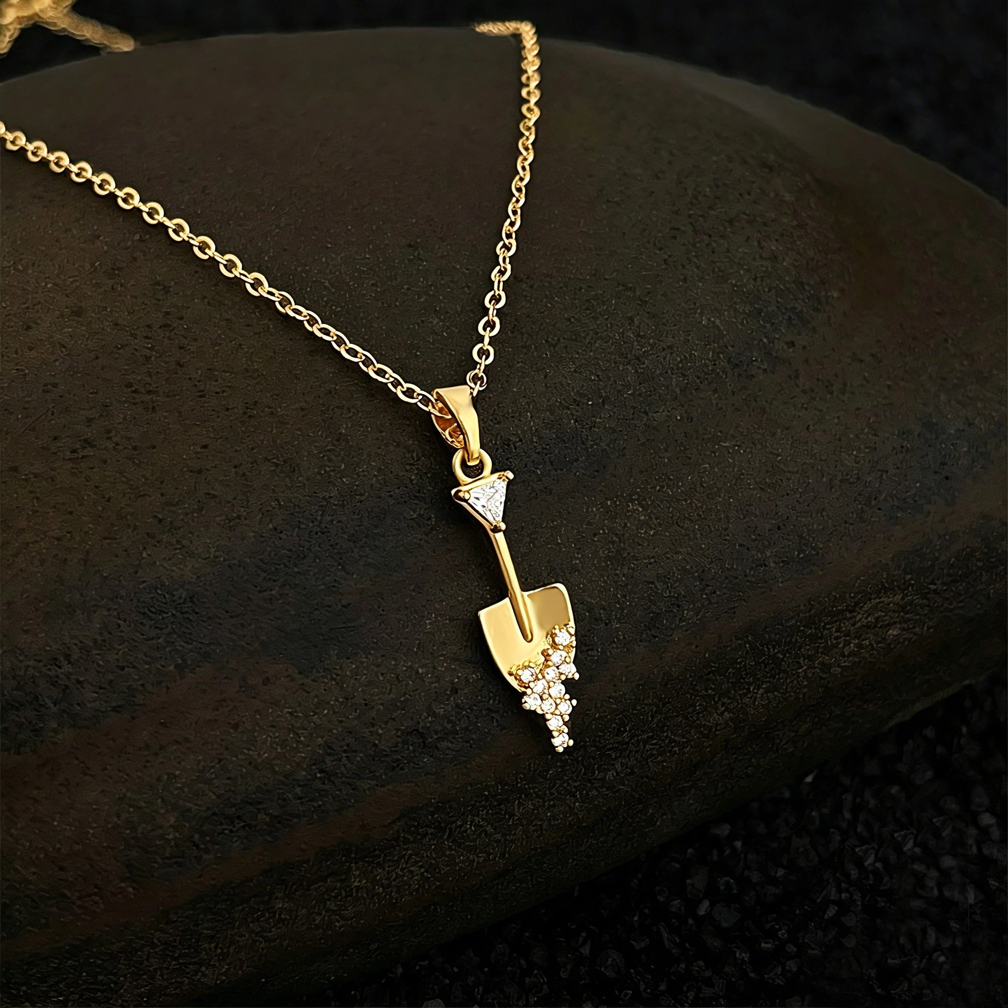 This is a necklace featuring a unique shovel-shaped pendant crafted with thick 18K gold plating, exuding a luxurious golden luster. The pendant is adorned with delicate micro-paved cubic zirconia stones, which sparkle brightly against the gold surface. Designed as a dainty pendant necklace, it showcases a sleek, minimalist style with intricate detailing on the shovel head and handle.