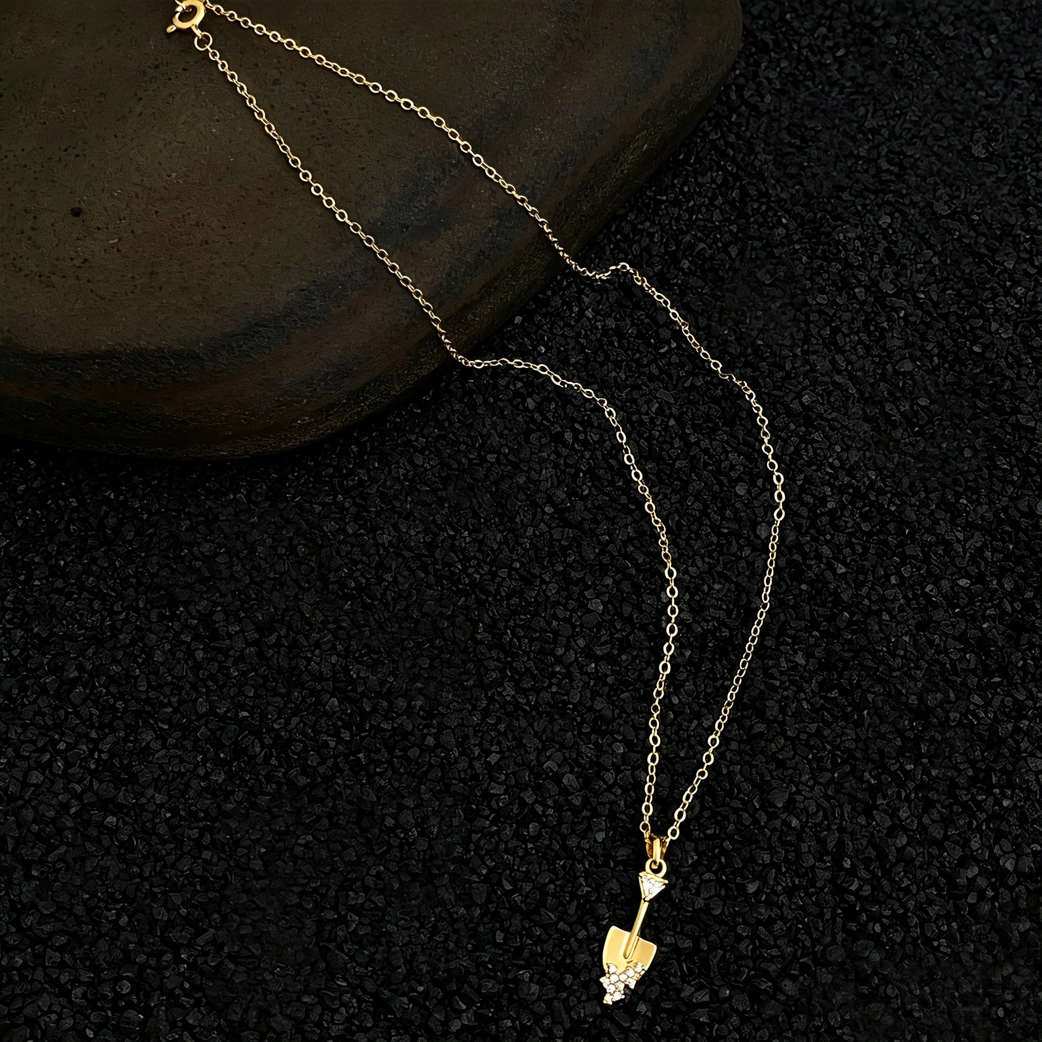 This is a necklace featuring a unique shovel-shaped pendant crafted with thick 18K gold plating, exuding a luxurious golden luster. The pendant is adorned with delicate micro-paved cubic zirconia stones, which sparkle brightly against the gold surface. Designed as a dainty pendant necklace, it showcases a sleek, minimalist style with intricate detailing on the shovel head and handle.