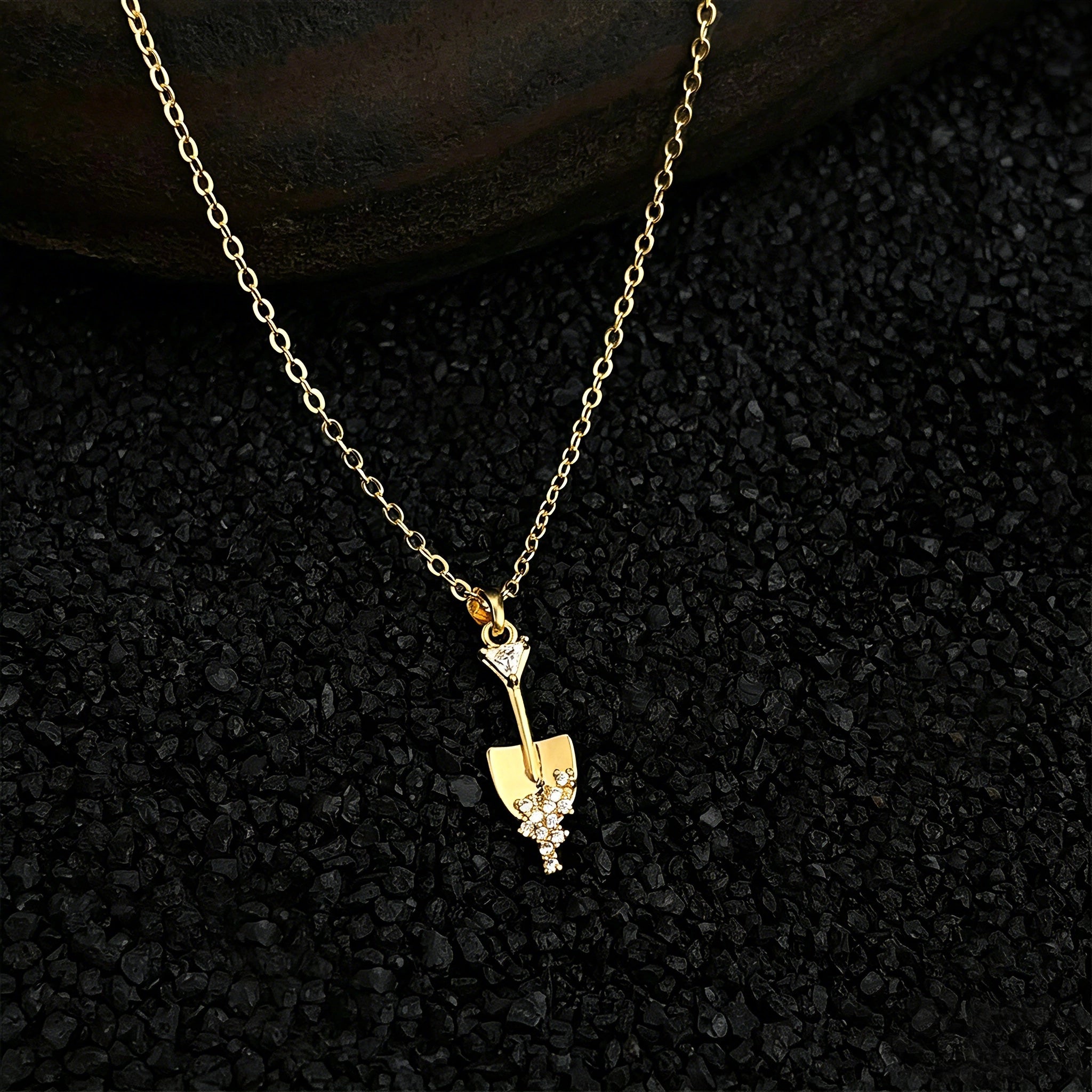This is a necklace featuring a unique shovel-shaped pendant crafted with thick 18K gold plating, exuding a luxurious golden luster. The pendant is adorned with delicate micro-paved cubic zirconia stones, which sparkle brightly against the gold surface. Designed as a dainty pendant necklace, it showcases a sleek, minimalist style with intricate detailing on the shovel head and handle.