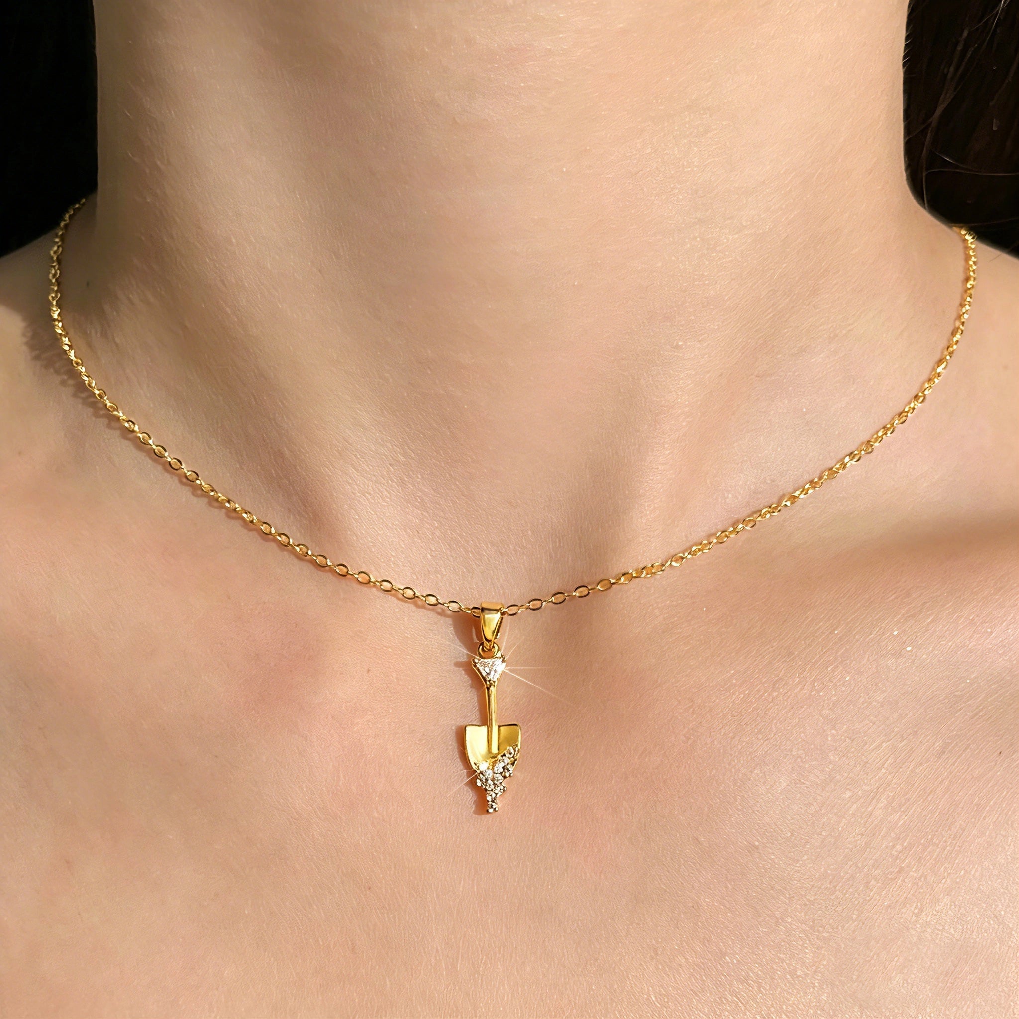 This is a necklace featuring a unique shovel-shaped pendant crafted with thick 18K gold plating, exuding a luxurious golden luster. The pendant is adorned with delicate micro-paved cubic zirconia stones, which sparkle brightly against the gold surface. Designed as a dainty pendant necklace, it showcases a sleek, minimalist style with intricate detailing on the shovel head and handle.