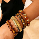 This 4-piece stackable bracelet set blends glossy tortoiseshell-pattern glass beads with polished stainless steel hardware. It includes flower-shaped, square, and oval beaded bracelets paired with a flexible gold-tone ribbed bangle. Perfect for layered styling, these warm earthy-toned accessories effortlessly elevate everyday and evening outfits.