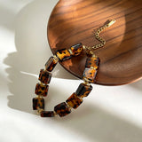This 4-piece stackable bracelet set blends glossy tortoiseshell-pattern glass beads with polished stainless steel hardware. It includes flower-shaped, square, and oval beaded bracelets paired with a flexible gold-tone ribbed bangle. Perfect for layered styling, these warm earthy-toned accessories effortlessly elevate everyday and evening outfits.