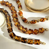 This 4-piece stackable bracelet set blends glossy tortoiseshell-pattern glass beads with polished stainless steel hardware. It includes flower-shaped, square, and oval beaded bracelets paired with a flexible gold-tone ribbed bangle. Perfect for layered styling, these warm earthy-toned accessories effortlessly elevate everyday and evening outfits.