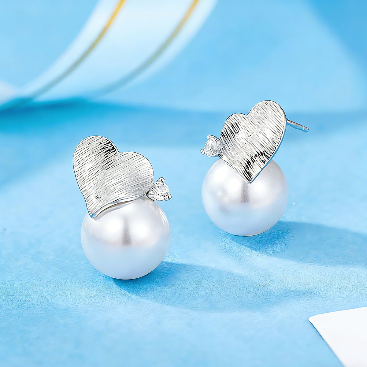 This minimalist pair of silver pearl stud earrings cleverly combine a chic silver heart-shaped pendant (11.7 mm wide) with a striking pearl teardrop (total height: 19.4 mm), with the pearl beneath the heart, creating a unique and stylish silhouette.