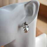 This minimalist pair of silver pearl stud earrings cleverly combine a chic silver heart-shaped pendant (11.7 mm wide) with a striking pearl teardrop (total height: 19.4 mm), with the pearl beneath the heart, creating a unique and stylish silhouette.