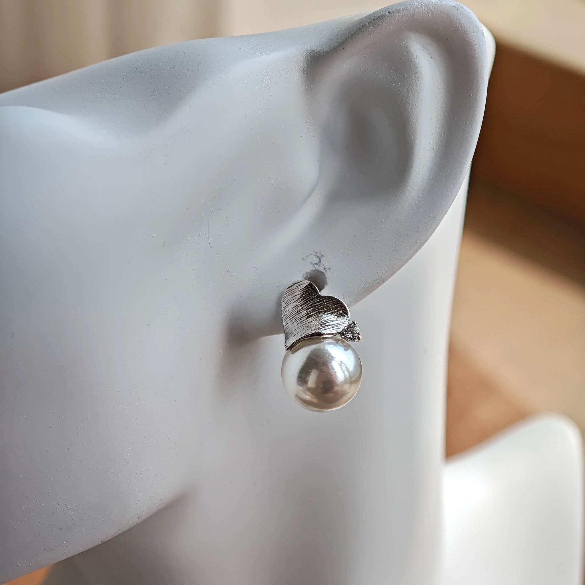 This minimalist pair of silver pearl stud earrings cleverly combine a chic silver heart-shaped pendant (11.7 mm wide) with a striking pearl teardrop (total height: 19.4 mm), with the pearl beneath the heart, creating a unique and stylish silhouette.