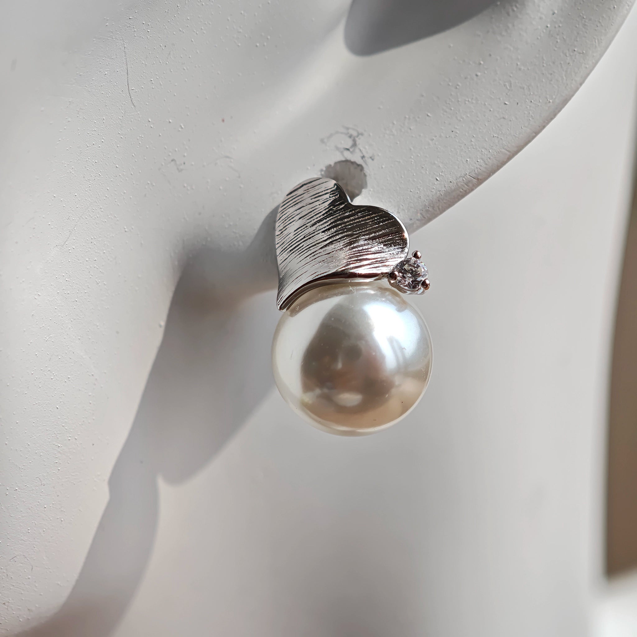 This minimalist pair of silver pearl stud earrings cleverly combine a chic silver heart-shaped pendant (11.7 mm wide) with a striking pearl teardrop (total height: 19.4 mm), with the pearl beneath the heart, creating a unique and stylish silhouette.