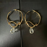 These artistic hoop earrings feature organic, knotted gold-toned metalwork that evokes a natural, twisted vine aesthetic. Suspended from each hoop is a freeform, clear charm with a textured, water-drop shape that catches light beautifully. The contrast between the warm, matte gold and the cool, translucent crystal creates a bold, avant-garde look.