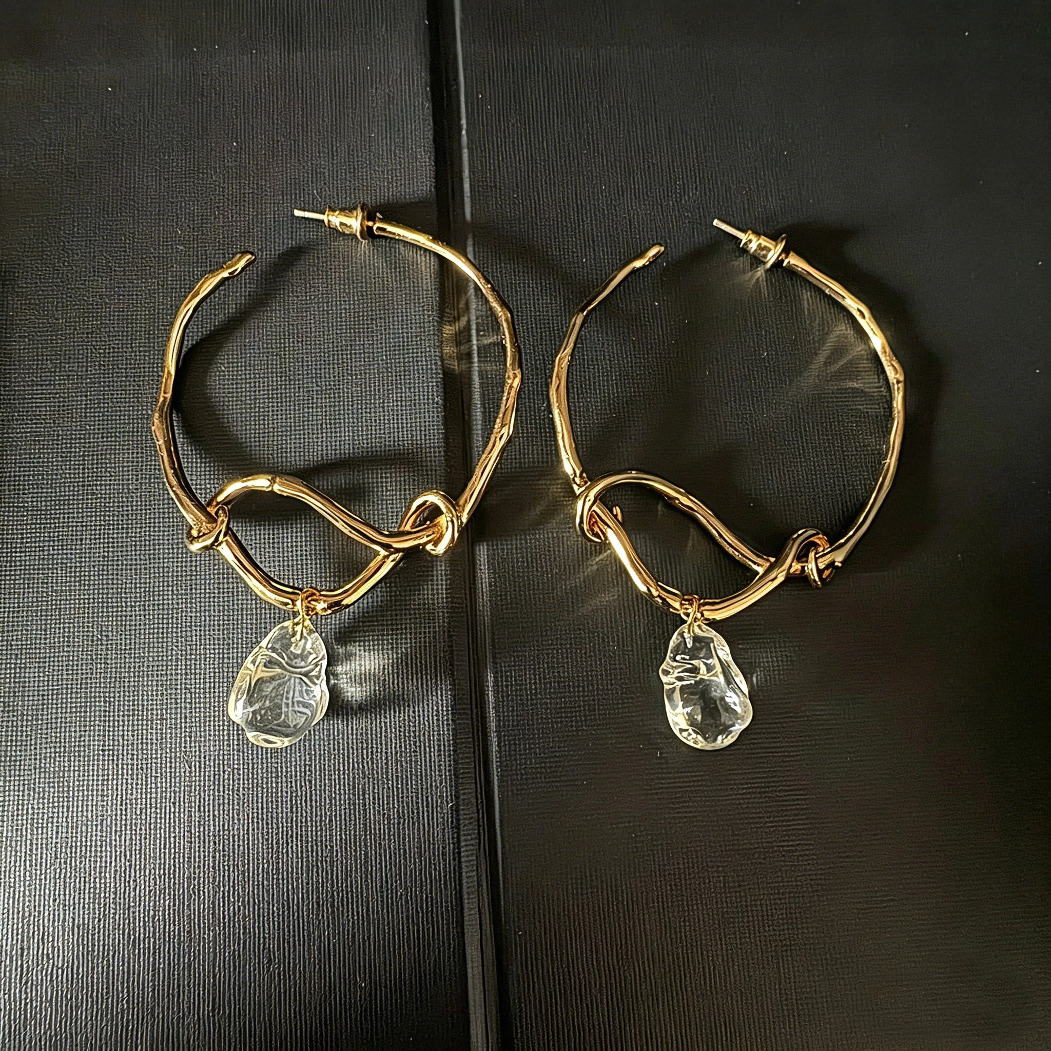 These artistic hoop earrings feature organic, knotted gold-toned metalwork that evokes a natural, twisted vine aesthetic. Suspended from each hoop is a freeform, clear charm with a textured, water-drop shape that catches light beautifully. The contrast between the warm, matte gold and the cool, translucent crystal creates a bold, avant-garde look.
