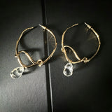 These artistic hoop earrings feature organic, knotted gold-toned metalwork that evokes a natural, twisted vine aesthetic. Suspended from each hoop is a freeform, clear charm with a textured, water-drop shape that catches light beautifully. The contrast between the warm, matte gold and the cool, translucent crystal creates a bold, avant-garde look.