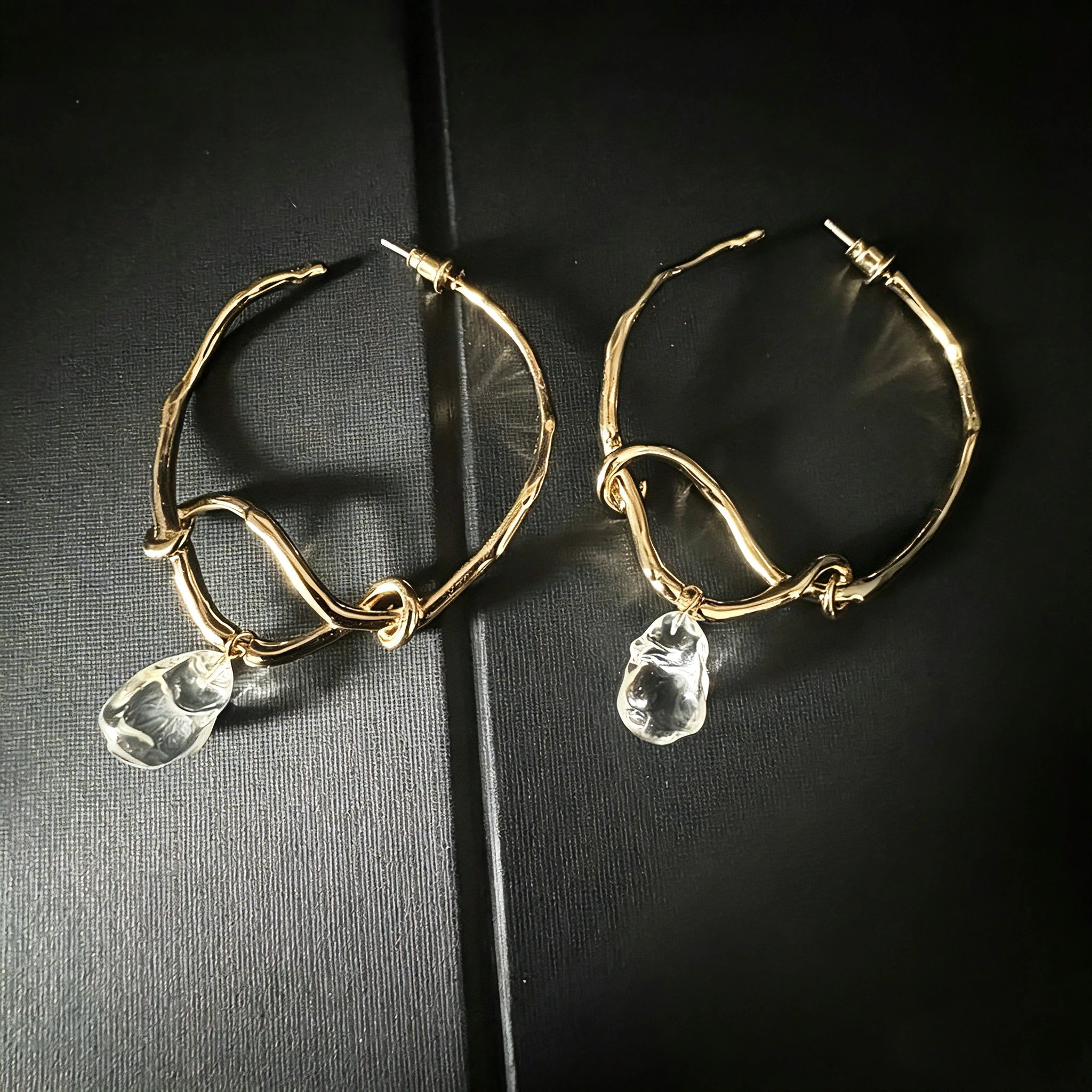 These artistic hoop earrings feature organic, knotted gold-toned metalwork that evokes a natural, twisted vine aesthetic. Suspended from each hoop is a freeform, clear charm with a textured, water-drop shape that catches light beautifully. The contrast between the warm, matte gold and the cool, translucent crystal creates a bold, avant-garde look.