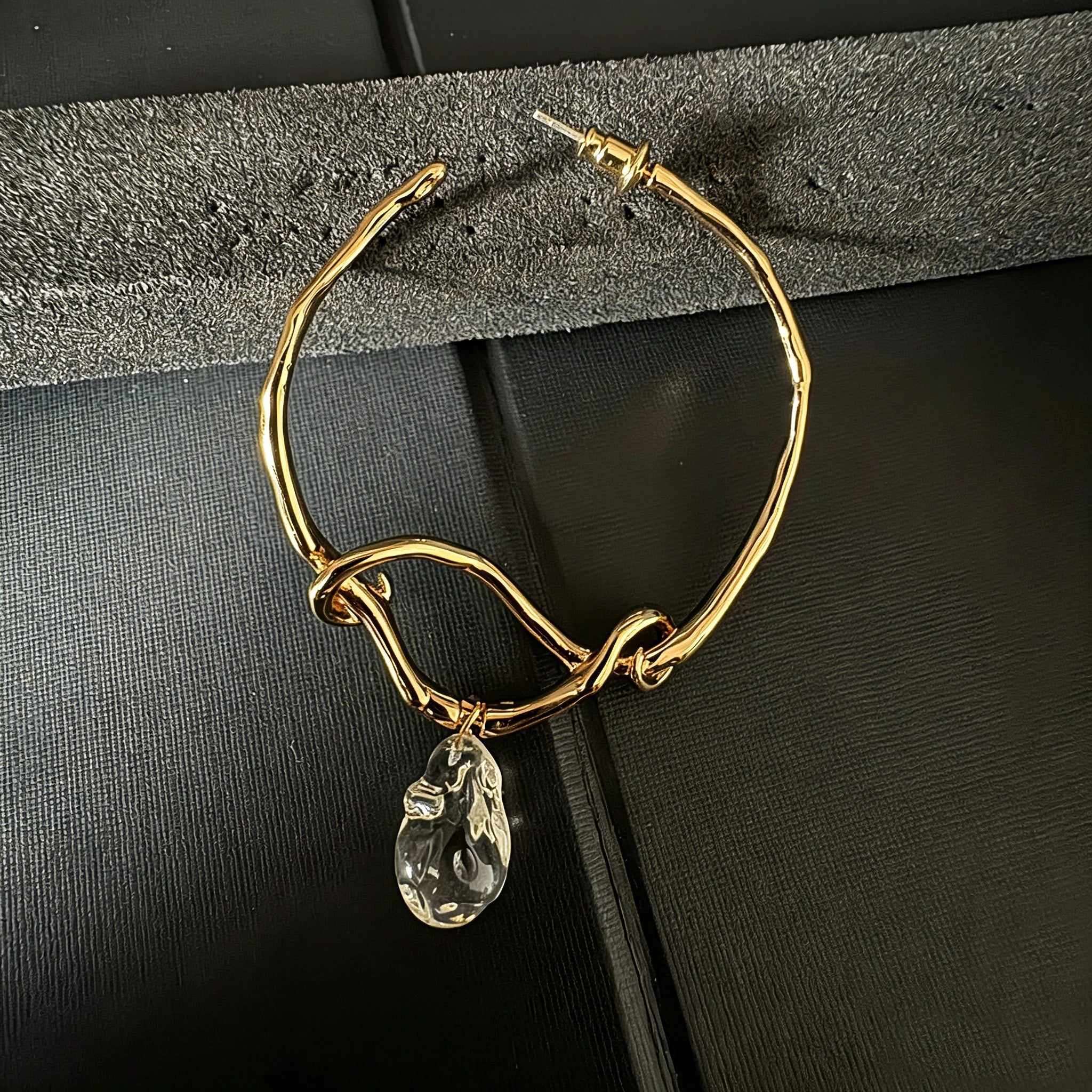 These artistic hoop earrings feature organic, knotted gold-toned metalwork that evokes a natural, twisted vine aesthetic. Suspended from each hoop is a freeform, clear charm with a textured, water-drop shape that catches light beautifully. The contrast between the warm, matte gold and the cool, translucent crystal creates a bold, avant-garde look.