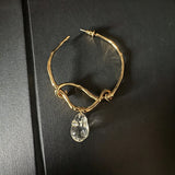 These artistic hoop earrings feature organic, knotted gold-toned metalwork that evokes a natural, twisted vine aesthetic. Suspended from each hoop is a freeform, clear charm with a textured, water-drop shape that catches light beautifully. The contrast between the warm, matte gold and the cool, translucent crystal creates a bold, avant-garde look.