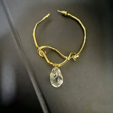 These artistic hoop earrings feature organic, knotted gold-toned metalwork that evokes a natural, twisted vine aesthetic. Suspended from each hoop is a freeform, clear charm with a textured, water-drop shape that catches light beautifully. The contrast between the warm, matte gold and the cool, translucent crystal creates a bold, avant-garde look.