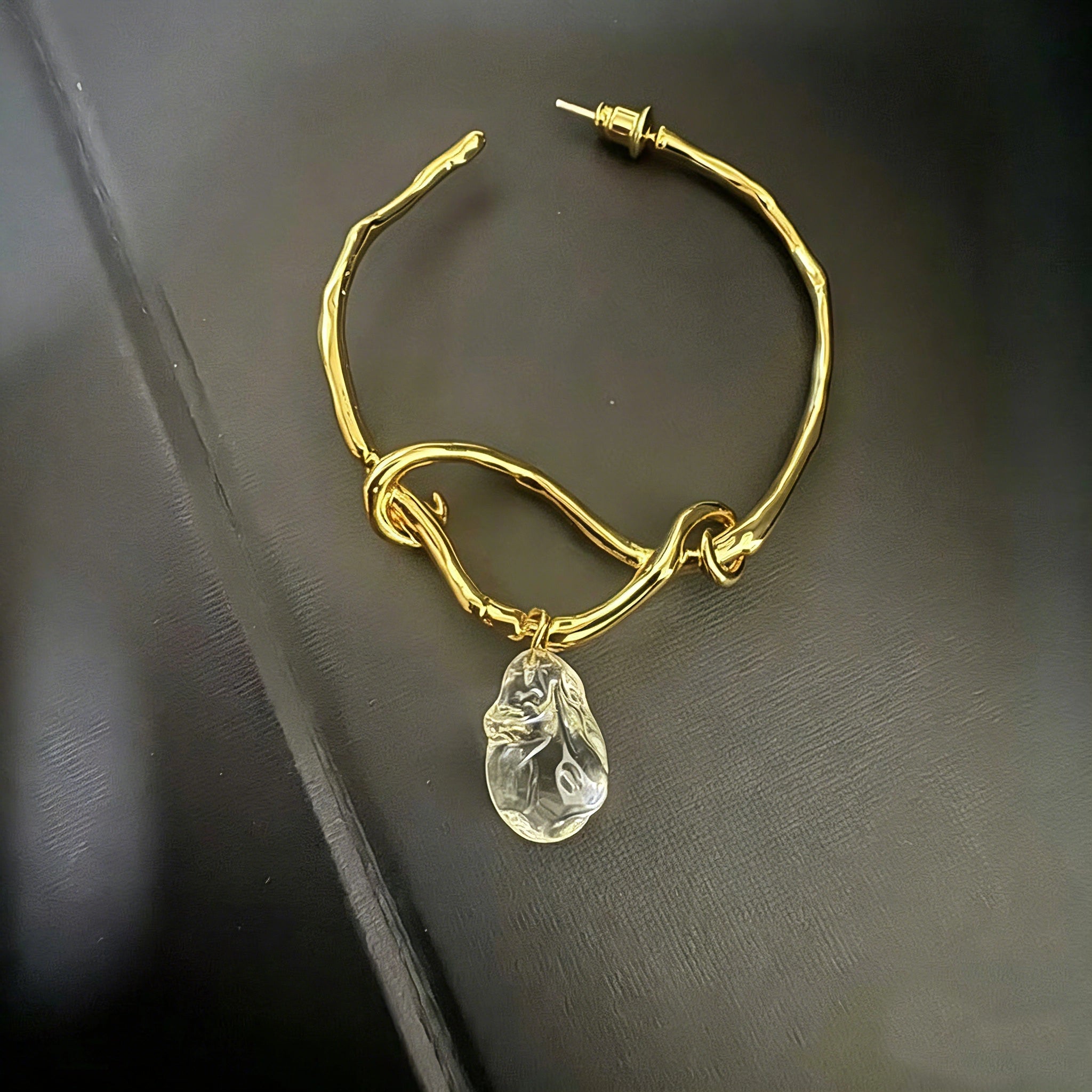These artistic hoop earrings feature organic, knotted gold-toned metalwork that evokes a natural, twisted vine aesthetic. Suspended from each hoop is a freeform, clear charm with a textured, water-drop shape that catches light beautifully. The contrast between the warm, matte gold and the cool, translucent crystal creates a bold, avant-garde look.