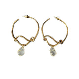 These artistic hoop earrings feature organic, knotted gold-toned metalwork that evokes a natural, twisted vine aesthetic. Suspended from each hoop is a freeform, clear charm with a textured, water-drop shape that catches light beautifully. The contrast between the warm, matte gold and the cool, translucent crystal creates a bold, avant-garde look.