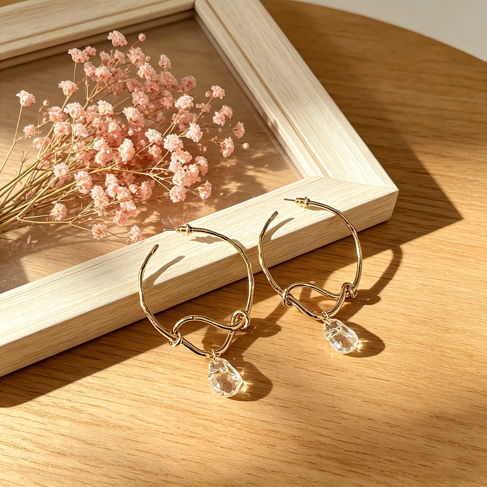These artistic hoop earrings feature organic, knotted gold-toned metalwork that evokes a natural, twisted vine aesthetic. Suspended from each hoop is a freeform, clear charm with a textured, water-drop shape that catches light beautifully. The contrast between the warm, matte gold and the cool, translucent crystal creates a bold, avant-garde look.