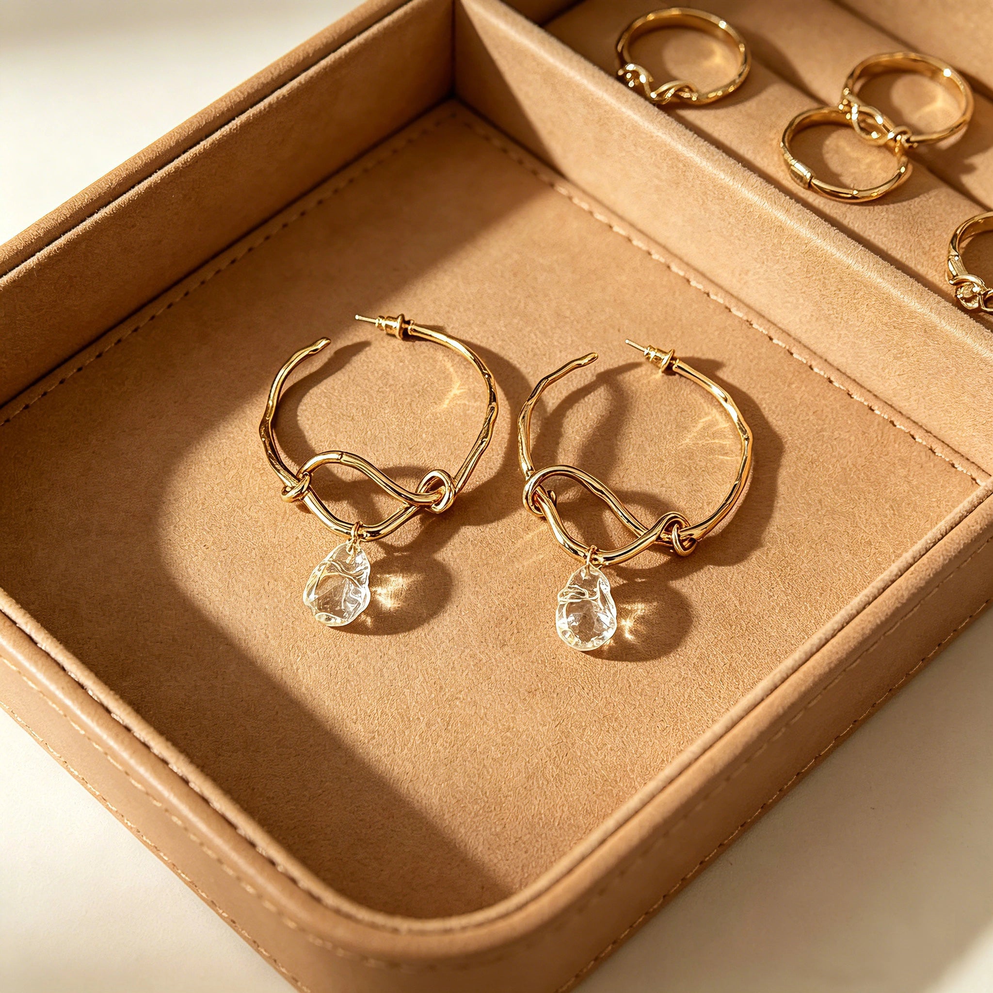 These artistic hoop earrings feature organic, knotted gold-toned metalwork that evokes a natural, twisted vine aesthetic. Suspended from each hoop is a freeform, clear charm with a textured, water-drop shape that catches light beautifully. The contrast between the warm, matte gold and the cool, translucent crystal creates a bold, avant-garde look.