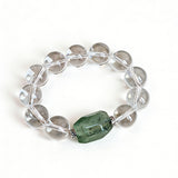 This stunning bracelet is crafted with a strand of perfectly round, high-clarity clear quartz beads, showcasing exceptional transparency and a brilliant, glass-like luster. At its center sits a striking free-form green gemstone accent, its natural inclusions and unique cut adding a one-of-a-kind, organic touch. The two stones are elegantly separated by delicate silver-toned decorative spacers, creating a balanced, sophisticated design that blends minimalist clarity with bold natural beauty.