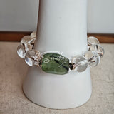 This stunning bracelet is crafted with a strand of perfectly round, high-clarity clear quartz beads, showcasing exceptional transparency and a brilliant, glass-like luster. At its center sits a striking free-form green gemstone accent, its natural inclusions and unique cut adding a one-of-a-kind, organic touch. The two stones are elegantly separated by delicate silver-toned decorative spacers, creating a balanced, sophisticated design that blends minimalist clarity with bold natural beauty.