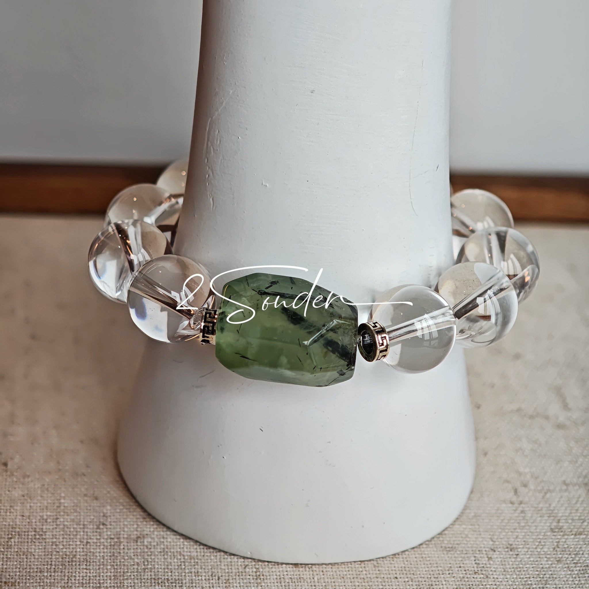 This stunning bracelet is crafted with a strand of perfectly round, high-clarity clear quartz beads, showcasing exceptional transparency and a brilliant, glass-like luster. At its center sits a striking free-form green gemstone accent, its natural inclusions and unique cut adding a one-of-a-kind, organic touch. The two stones are elegantly separated by delicate silver-toned decorative spacers, creating a balanced, sophisticated design that blends minimalist clarity with bold natural beauty.
