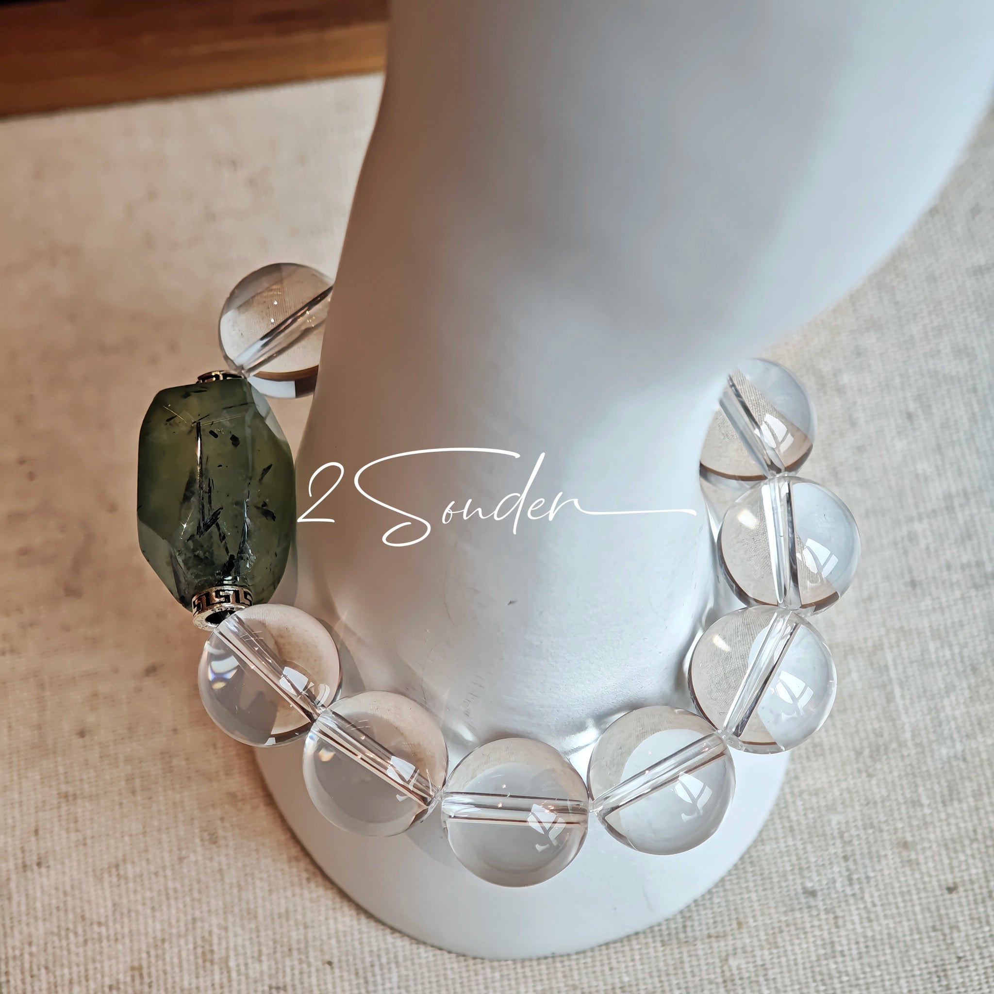 This stunning bracelet is crafted with a strand of perfectly round, high-clarity clear quartz beads, showcasing exceptional transparency and a brilliant, glass-like luster. At its center sits a striking free-form green gemstone accent, its natural inclusions and unique cut adding a one-of-a-kind, organic touch. The two stones are elegantly separated by delicate silver-toned decorative spacers, creating a balanced, sophisticated design that blends minimalist clarity with bold natural beauty.