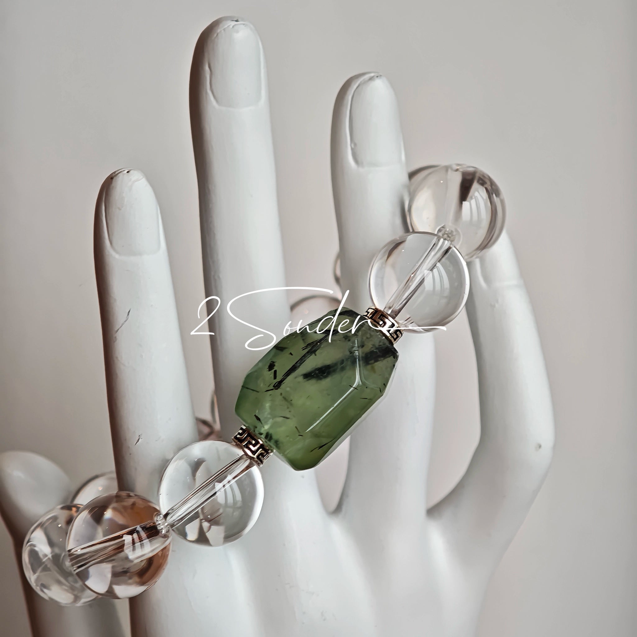 This stunning bracelet is crafted with a strand of perfectly round, high-clarity clear quartz beads, showcasing exceptional transparency and a brilliant, glass-like luster. At its center sits a striking free-form green gemstone accent, its natural inclusions and unique cut adding a one-of-a-kind, organic touch. The two stones are elegantly separated by delicate silver-toned decorative spacers, creating a balanced, sophisticated design that blends minimalist clarity with bold natural beauty.