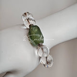 This stunning bracelet is crafted with a strand of perfectly round, high-clarity clear quartz beads, showcasing exceptional transparency and a brilliant, glass-like luster. At its center sits a striking free-form green gemstone accent, its natural inclusions and unique cut adding a one-of-a-kind, organic touch. The two stones are elegantly separated by delicate silver-toned decorative spacers, creating a balanced, sophisticated design that blends minimalist clarity with bold natural beauty.