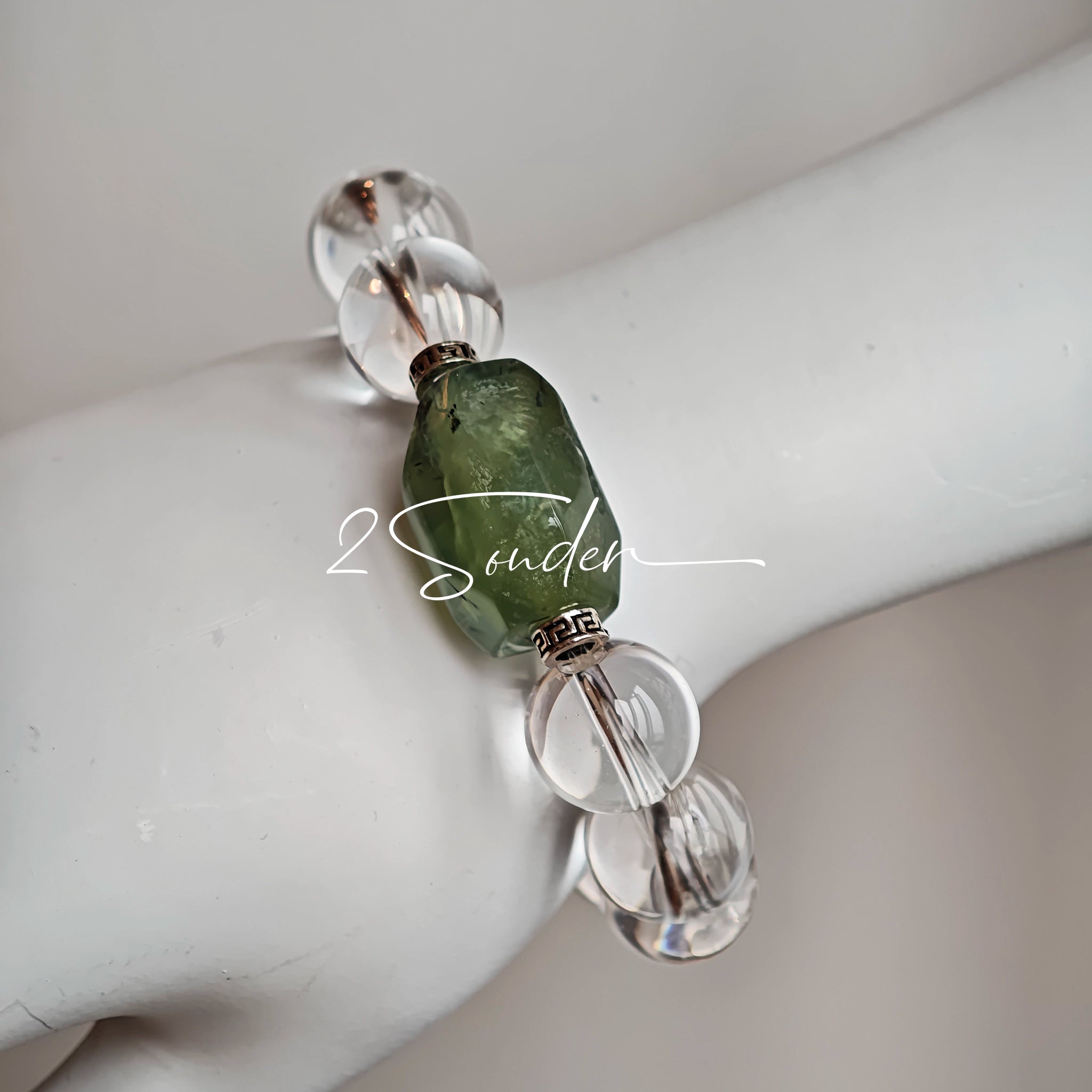 This stunning bracelet is crafted with a strand of perfectly round, high-clarity clear quartz beads, showcasing exceptional transparency and a brilliant, glass-like luster. At its center sits a striking free-form green gemstone accent, its natural inclusions and unique cut adding a one-of-a-kind, organic touch. The two stones are elegantly separated by delicate silver-toned decorative spacers, creating a balanced, sophisticated design that blends minimalist clarity with bold natural beauty.