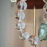 This stunning bracelet is crafted with a strand of perfectly round, high-clarity clear quartz beads, showcasing exceptional transparency and a brilliant, glass-like luster. At its center sits a striking free-form green gemstone accent, its natural inclusions and unique cut adding a one-of-a-kind, organic touch. The two stones are elegantly separated by delicate silver-toned decorative spacers, creating a balanced, sophisticated design that blends minimalist clarity with bold natural beauty.