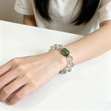 This stunning bracelet is crafted with a strand of perfectly round, high-clarity clear quartz beads, showcasing exceptional transparency and a brilliant, glass-like luster. At its center sits a striking free-form green gemstone accent, its natural inclusions and unique cut adding a one-of-a-kind, organic touch. The two stones are elegantly separated by delicate silver-toned decorative spacers, creating a balanced, sophisticated design that blends minimalist clarity with bold natural beauty.