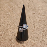 This striking bypass ring showcases two large, brilliant cushion-cut clear stones held securely in classic four-prong settings. The elegant twisted band is lined with a row of sparkling accent Cubic Zirconia stones, delivering a high-end, light luxury aesthetic that instantly catches the eye.