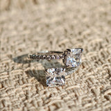 This striking bypass ring showcases two large, brilliant cushion-cut clear stones held securely in classic four-prong settings. The elegant twisted band is lined with a row of sparkling accent Cubic Zirconia stones, delivering a high-end, light luxury aesthetic that instantly catches the eye.
