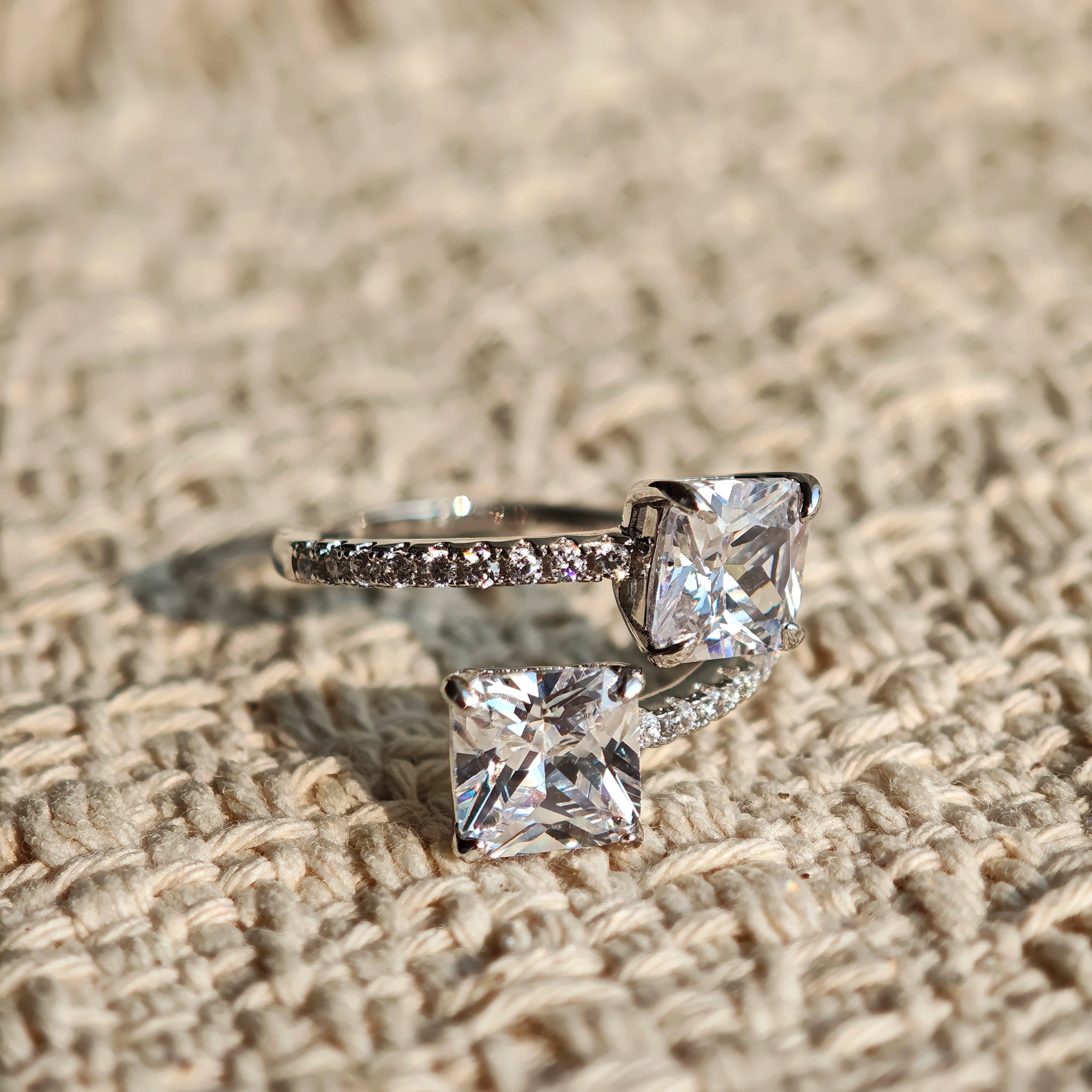 This striking bypass ring showcases two large, brilliant cushion-cut clear stones held securely in classic four-prong settings. The elegant twisted band is lined with a row of sparkling accent Cubic Zirconia stones, delivering a high-end, light luxury aesthetic that instantly catches the eye.