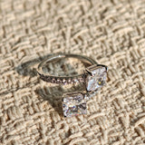 This striking bypass ring showcases two large, brilliant cushion-cut clear stones held securely in classic four-prong settings. The elegant twisted band is lined with a row of sparkling accent Cubic Zirconia stones, delivering a high-end, light luxury aesthetic that instantly catches the eye.