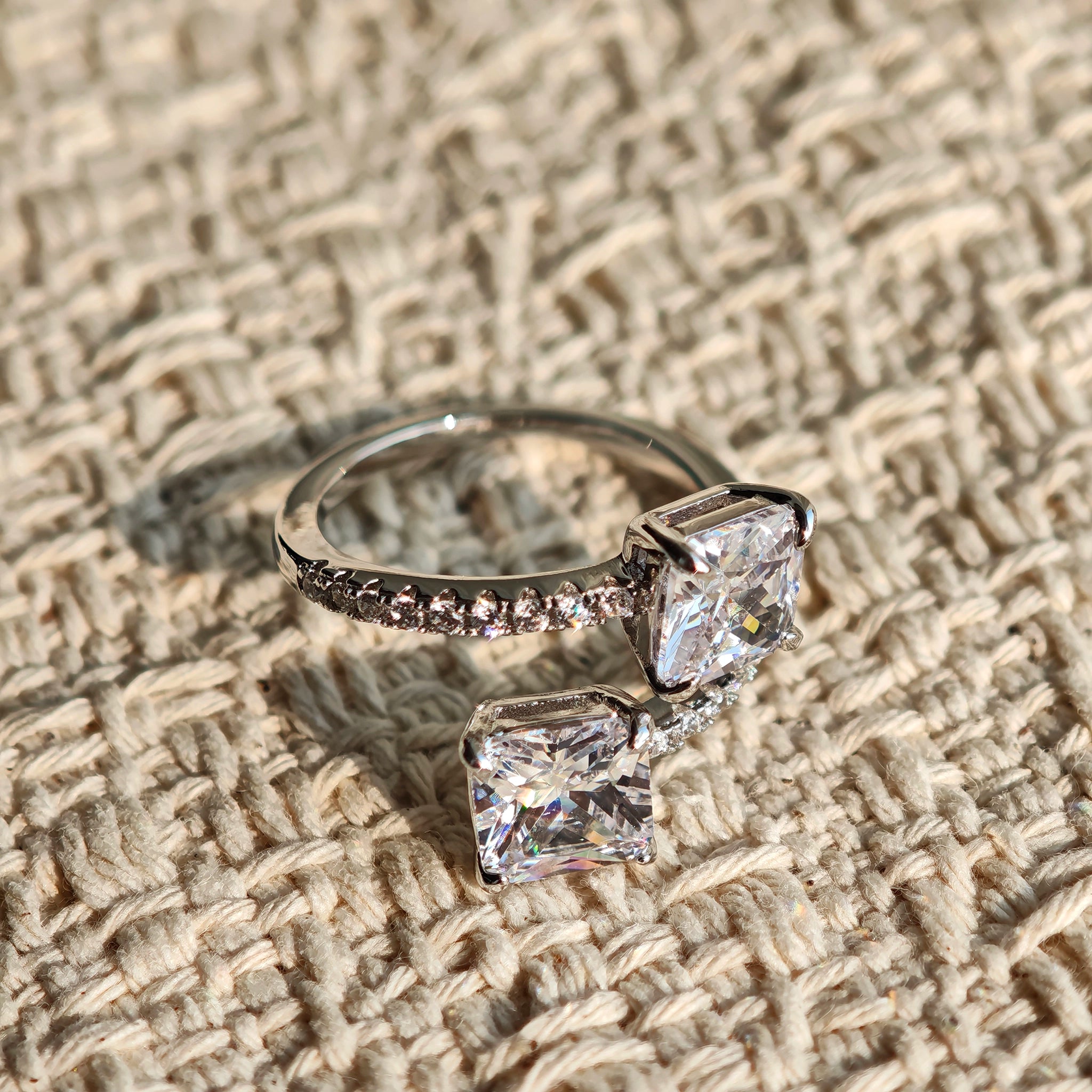 This striking bypass ring showcases two large, brilliant cushion-cut clear stones held securely in classic four-prong settings. The elegant twisted band is lined with a row of sparkling accent Cubic Zirconia stones, delivering a high-end, light luxury aesthetic that instantly catches the eye.