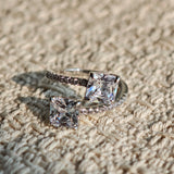 This striking bypass ring showcases two large, brilliant cushion-cut clear stones held securely in classic four-prong settings. The elegant twisted band is lined with a row of sparkling accent Cubic Zirconia stones, delivering a high-end, light luxury aesthetic that instantly catches the eye.