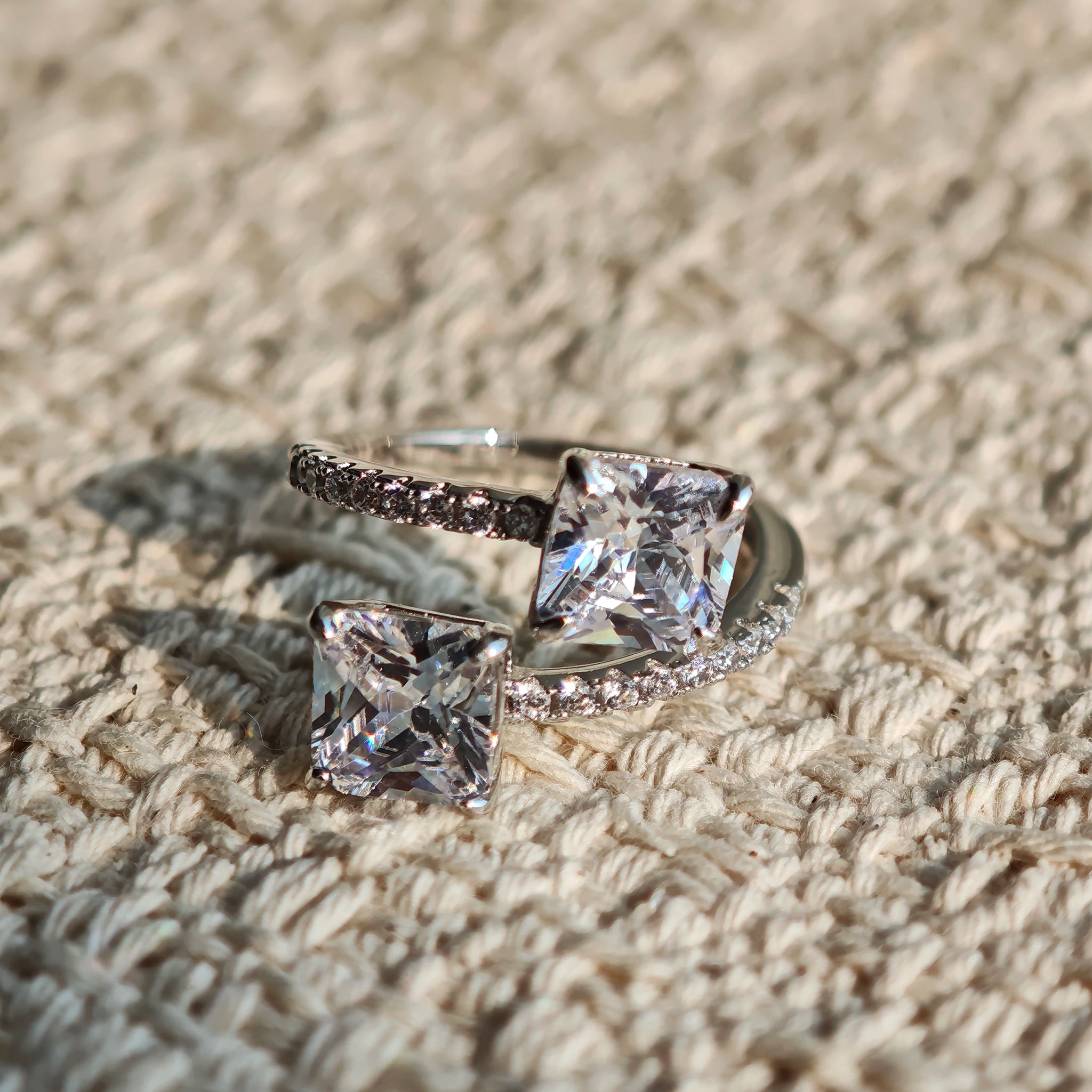 This striking bypass ring showcases two large, brilliant cushion-cut clear stones held securely in classic four-prong settings. The elegant twisted band is lined with a row of sparkling accent Cubic Zirconia stones, delivering a high-end, light luxury aesthetic that instantly catches the eye.
